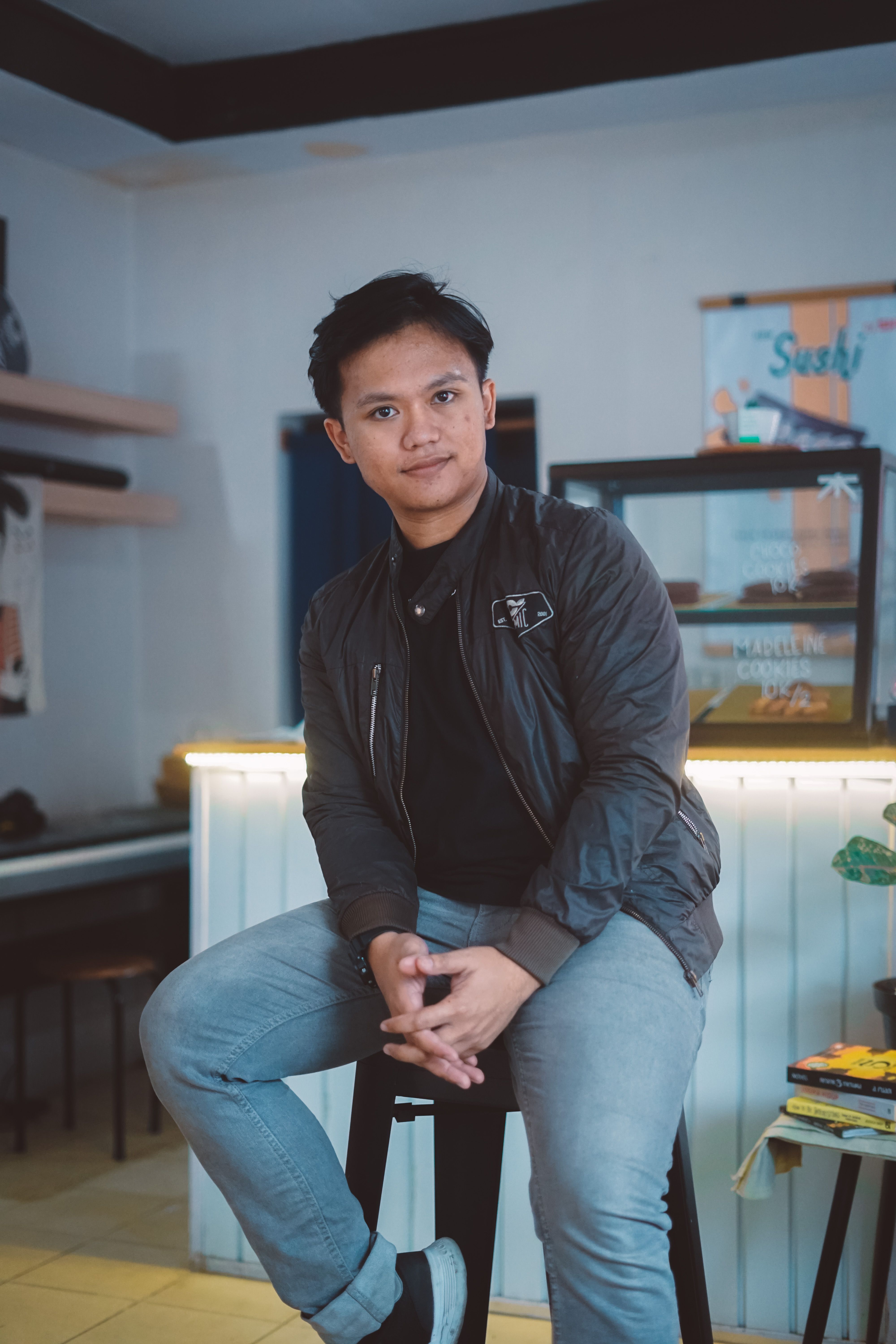 Man sitting casually on a stool in a cafe, wearing a jacket and jeans, with a sushi poster in the background.