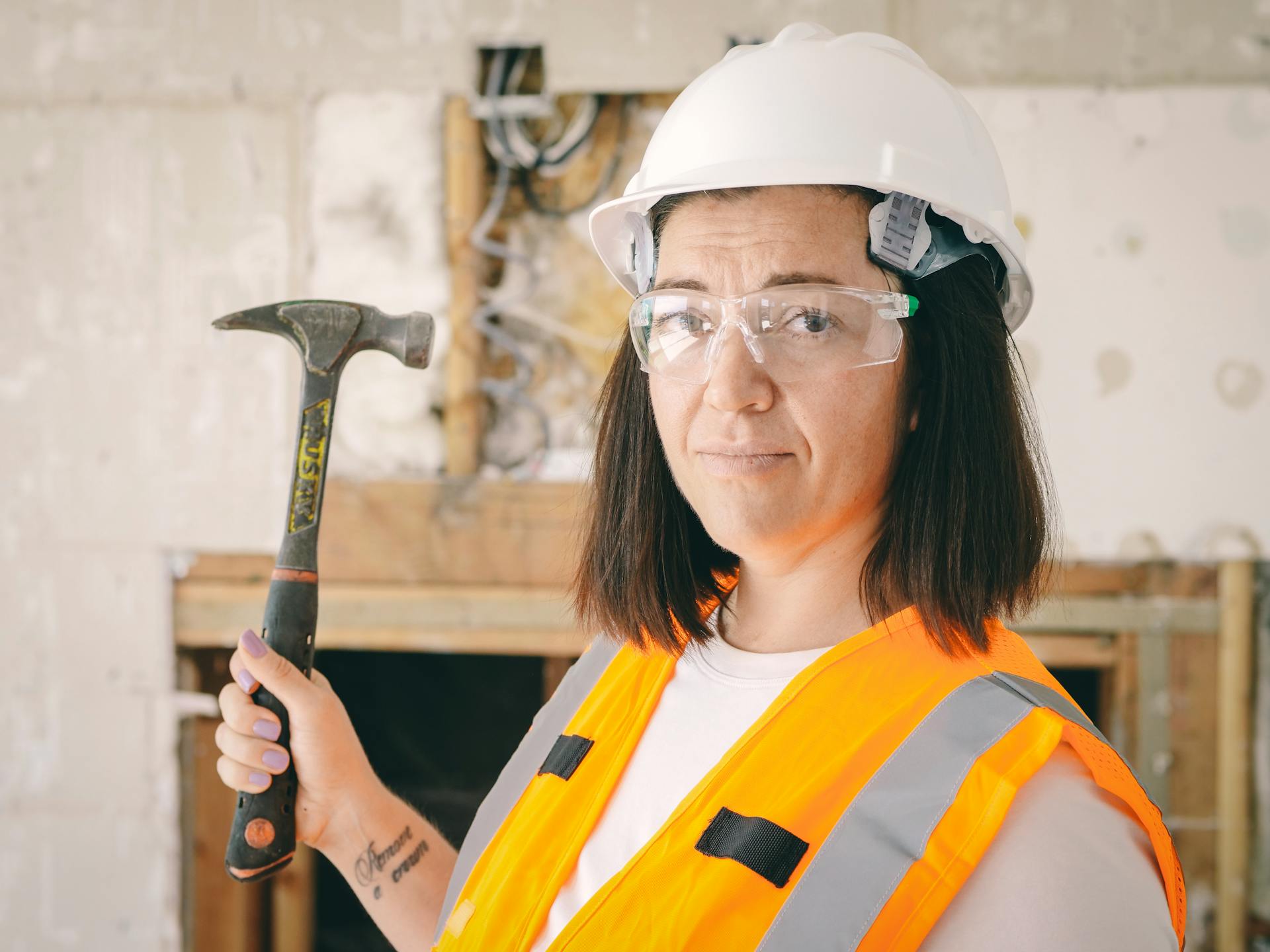 Confident female construction worker holding a hammer, equipped with safety gear including a hard hat and reflective vest.