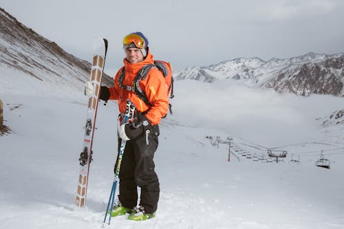 Mann, Der Orange Und Schwarzen Schneeanzug Mit Ski Set Auf Schnee In Der Nähe Von Seilbahnen Trägt