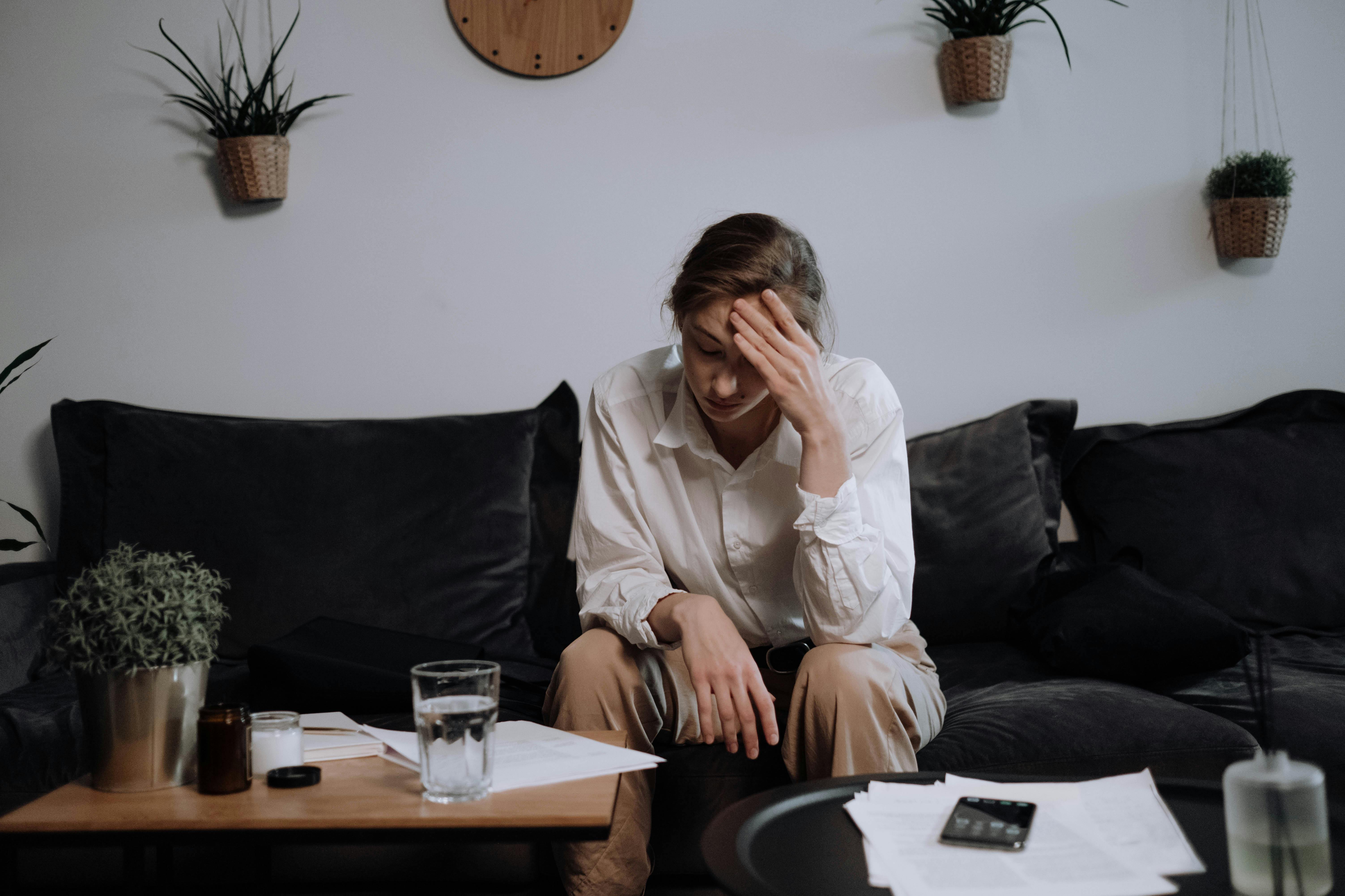 Woman experiencing stress and headache while sitting on a sofa at home, surrounded by papers