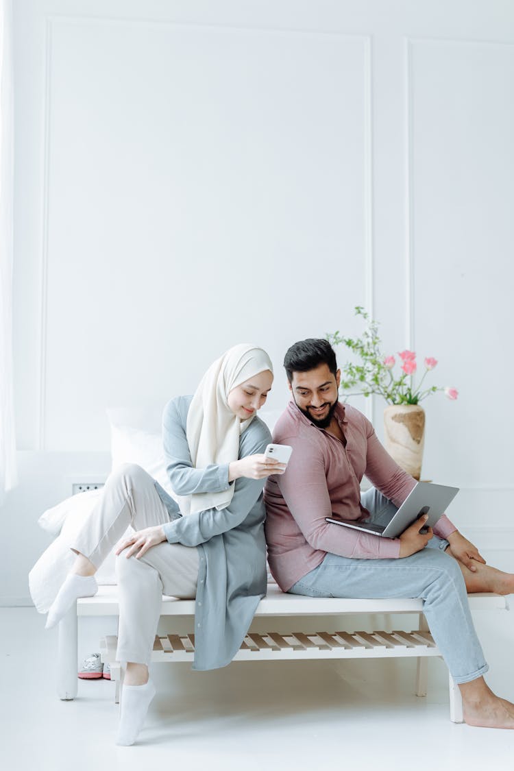 Couple Sitting On White Bedroom Bench With Cellphone And Laptop