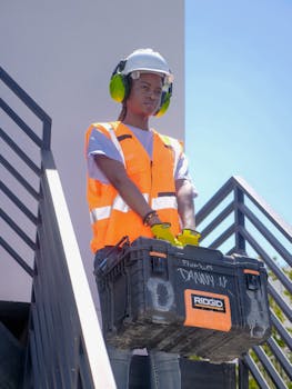 Photo by Kindel Media Black female construction worker wearing PPE holds a toolbox on stairs outdoors.
