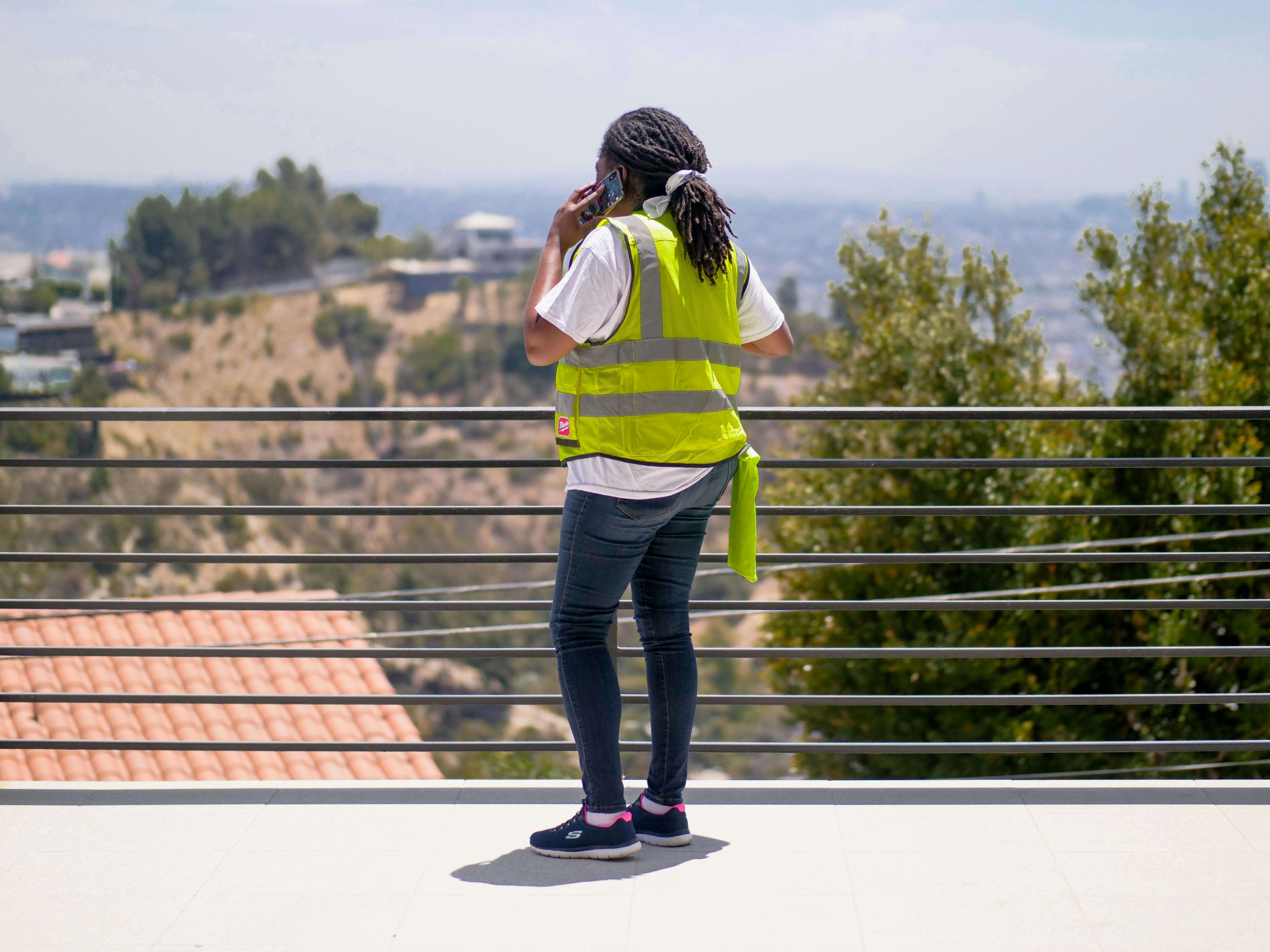 A construction worker wearing PPE and a reflective vest, talking on the phone outdoors.