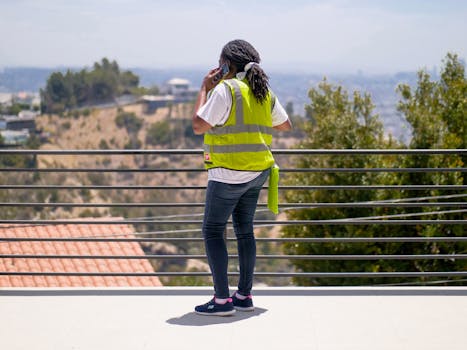A construction worker wearing PPE and a reflective vest, talking on the phone outdoors.