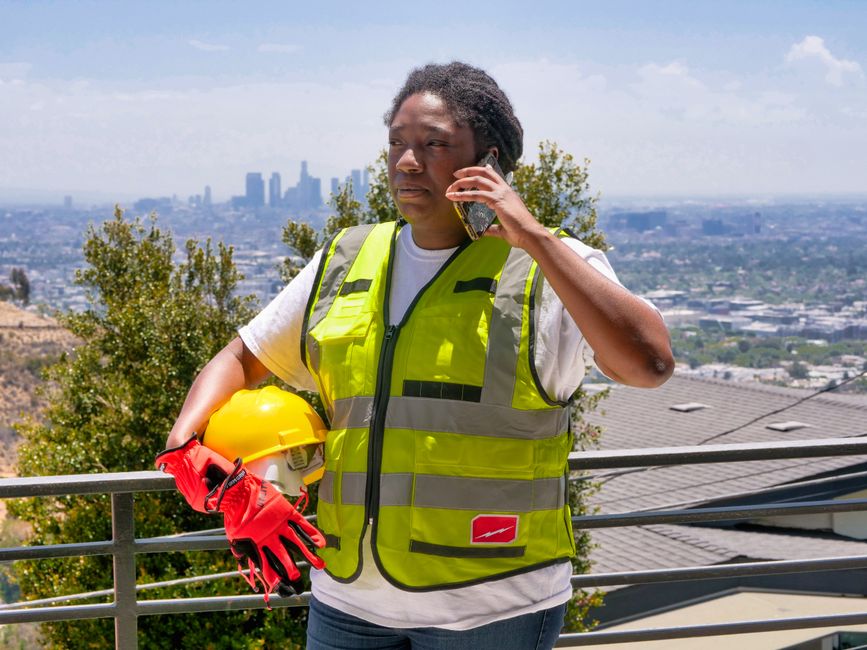 Black woman in safety vest and hardhat holding phone with cityscape background.