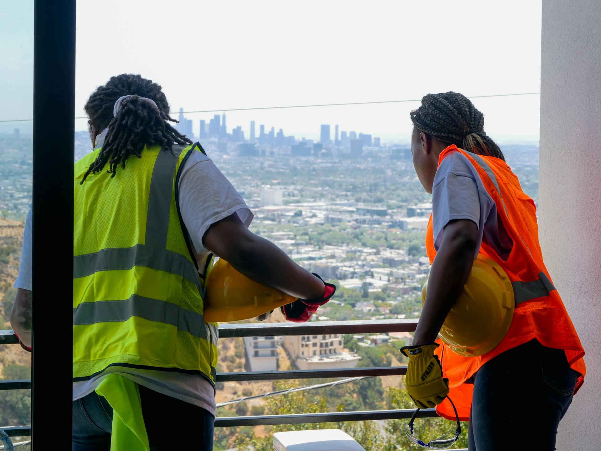 Construction workers on a balcony viewing a city skyline, wearing safety gear.