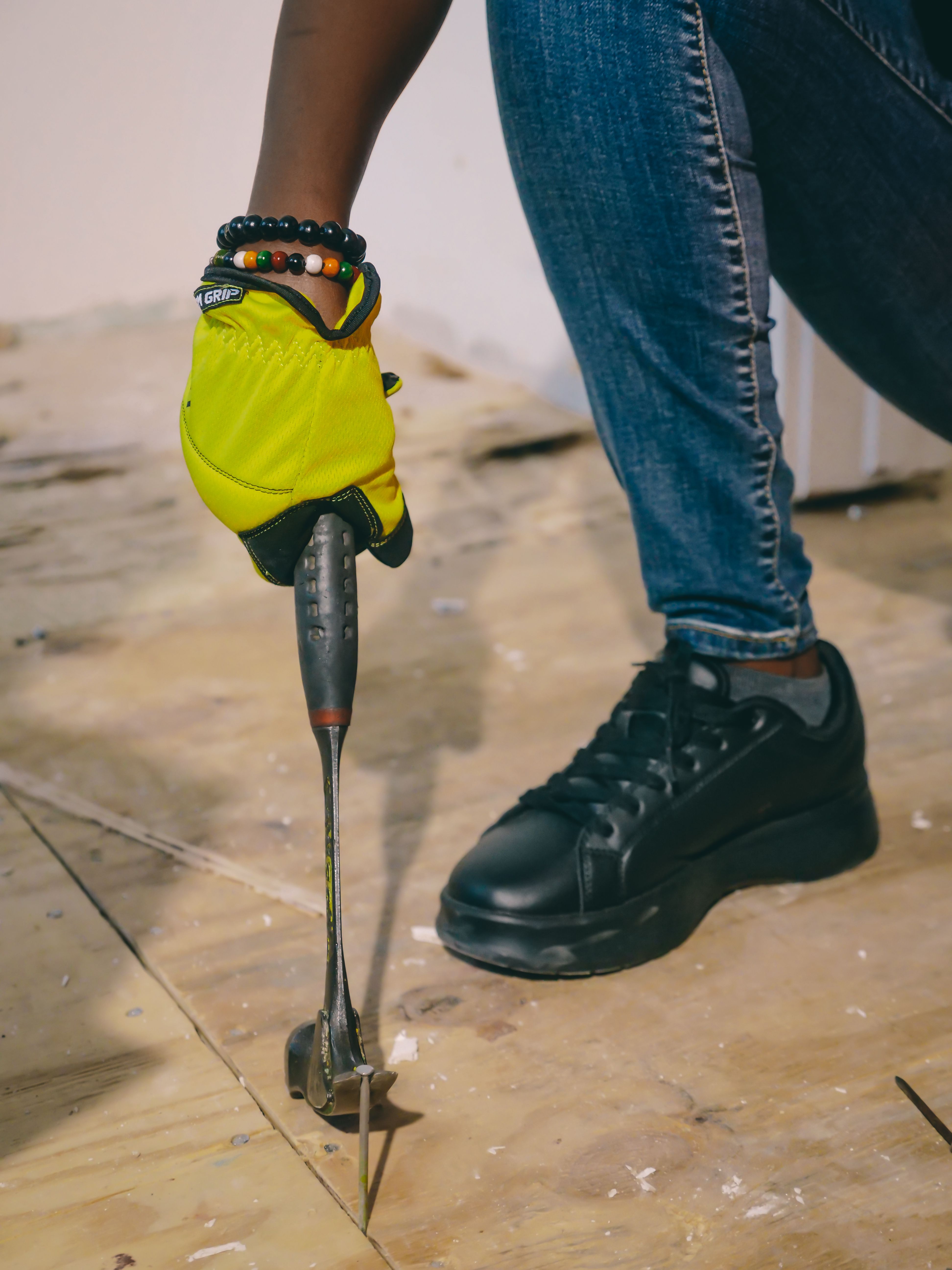 A gloved hand levering a nail out of a bare plywood subfloor with a pry bar, the worker's boot braced alongside