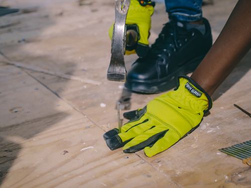 Free A worker wearing bright gloves uses a hammer on a plywood surface, emphasizing safety and construction. Stock Photo