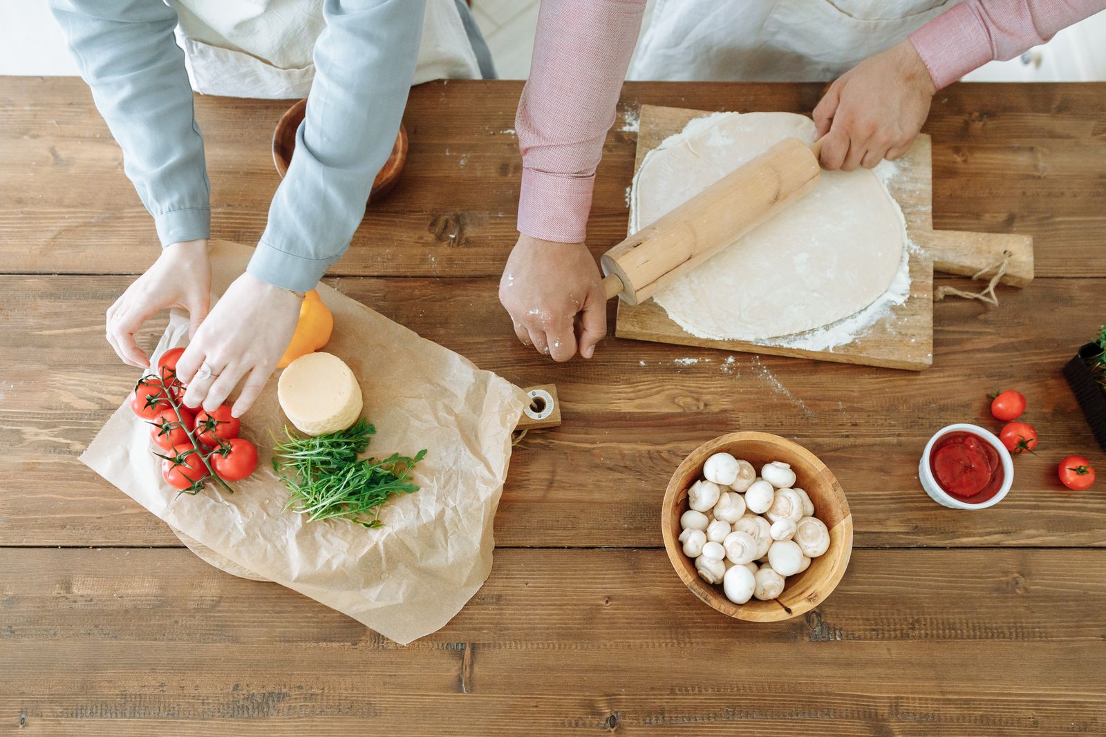 Couple rolling pizza dough and arranging fresh ingredients on wooden table, creating a homemade meal.