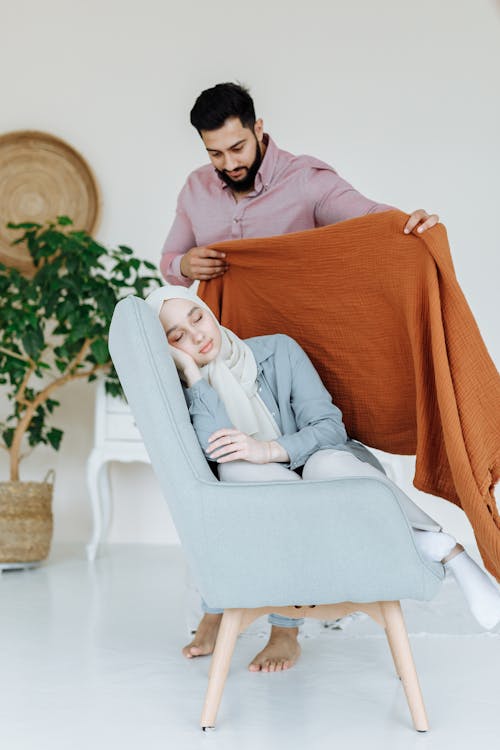 A Man in Pink Long Sleeves Putting  Blanket on the Woman Sleeping on the Chair