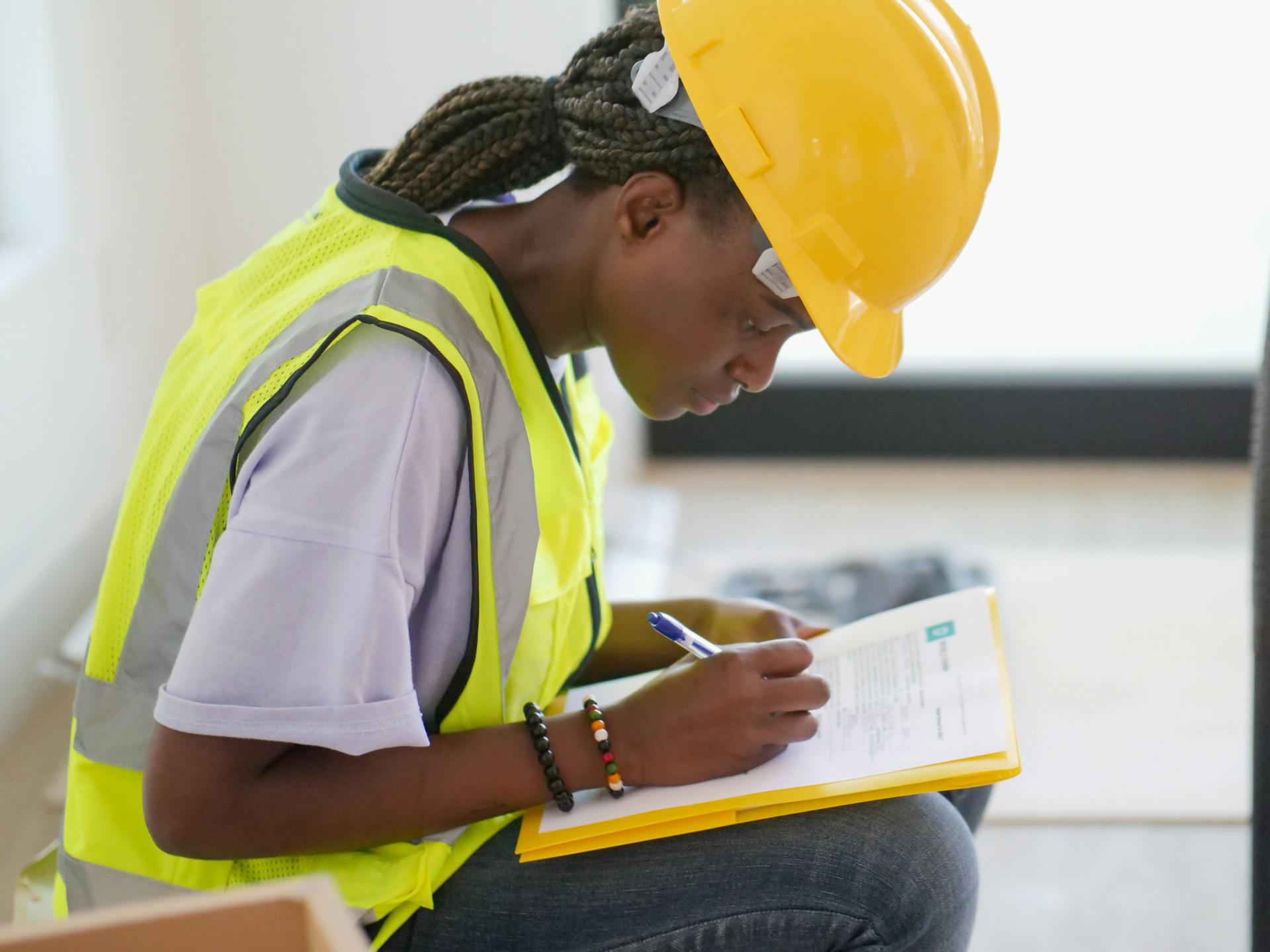 Focused female engineer in a safety helmet and vest writing on a clipboard indoors.