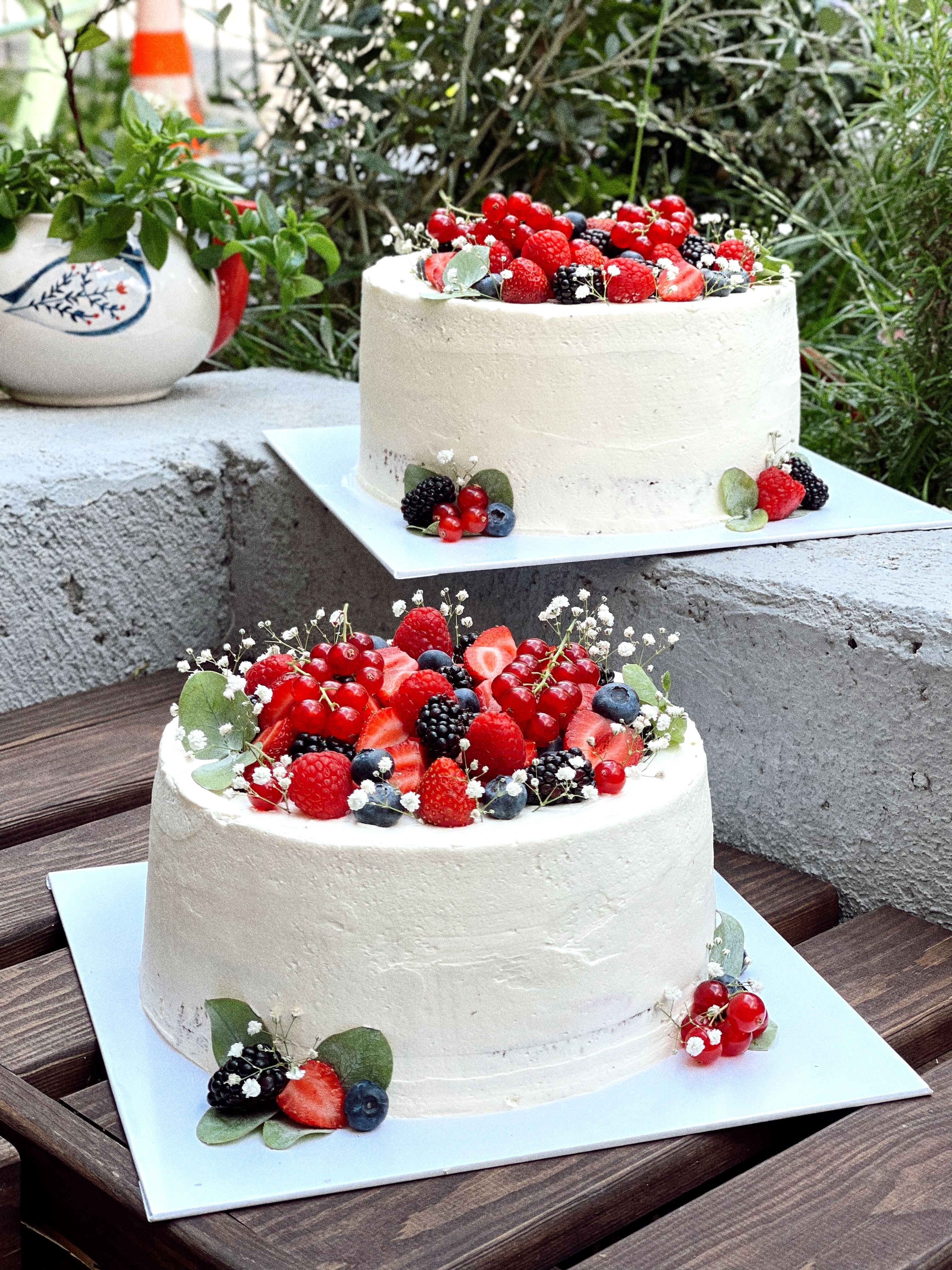 Two elegant white cakes adorned with fresh berries and flowers, set outdoors.