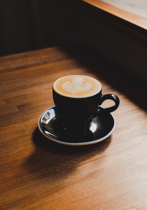 Free A latte with intricate foam art in a black cup on a wooden table by the window. Stock Photo