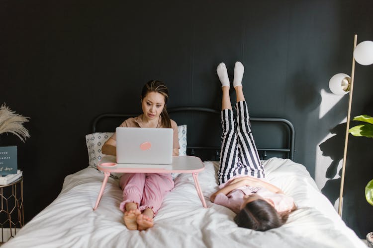 A Mother Lying On Bed With Her Daughter While Working