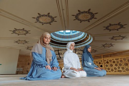 Free Three Muslim women in traditional attire praying inside a beautifully decorated mosque with intricate ceiling designs. Stock Photo