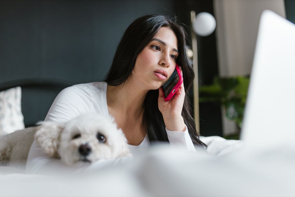 Woman relaxing on bed with her dog, talking on a smartphone indoors.