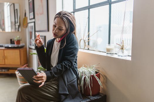 Asian woman multitasks at home using a tablet and phone in a cozy setting.