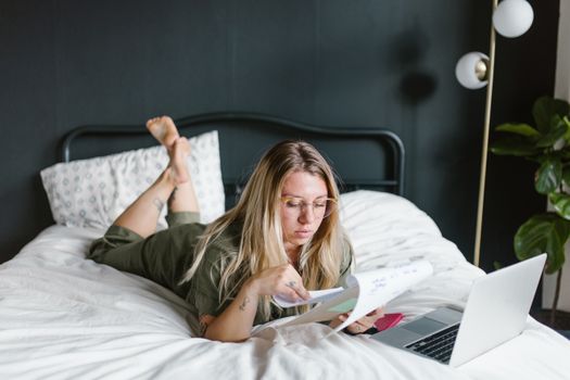 Young woman working from home, lying on bed with laptop and documents.
