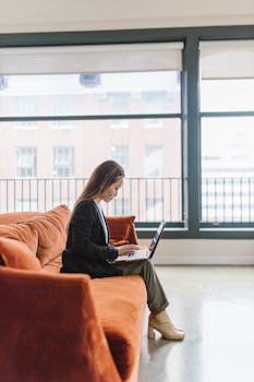 A woman working remotely on a laptop while sitting on an orange sofa in a modern living room.