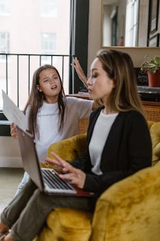 A mother working from home on a laptop while her daughter interacts beside her.