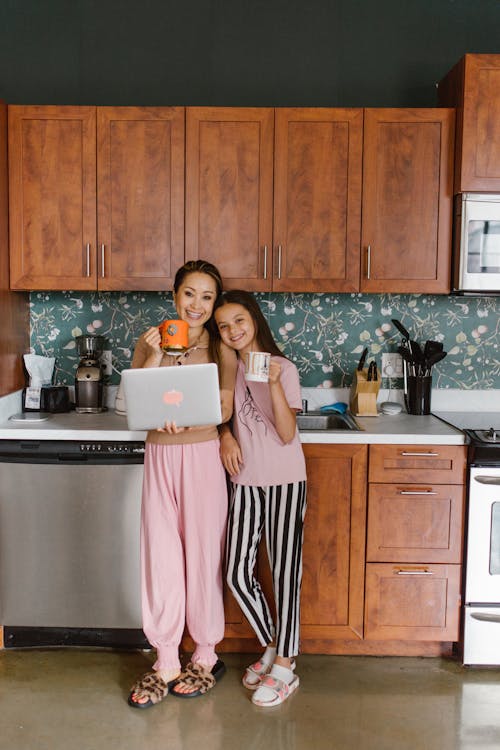 Free A Woman Holding Her Laptop while Standing Beside Her Daughter in the Kitchen Stock Photo