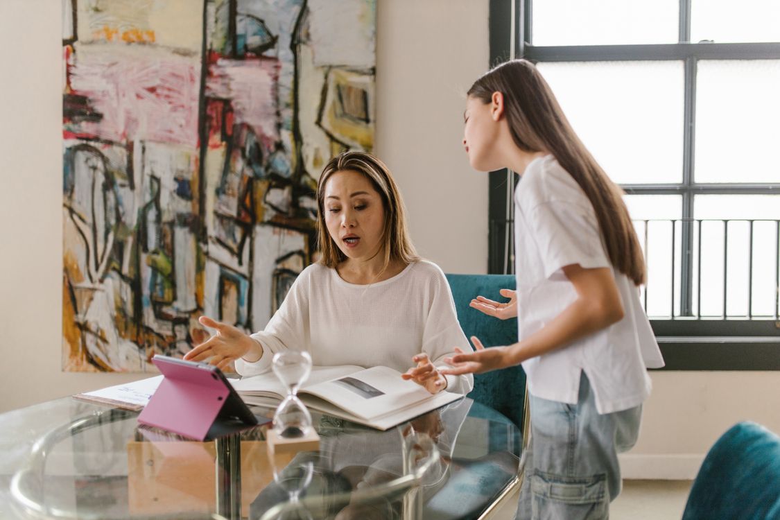A mother and teen daughter have an open conversation at a desk with a tablet nearby, representing cyberbullying awareness and parent-child digital safety discussions