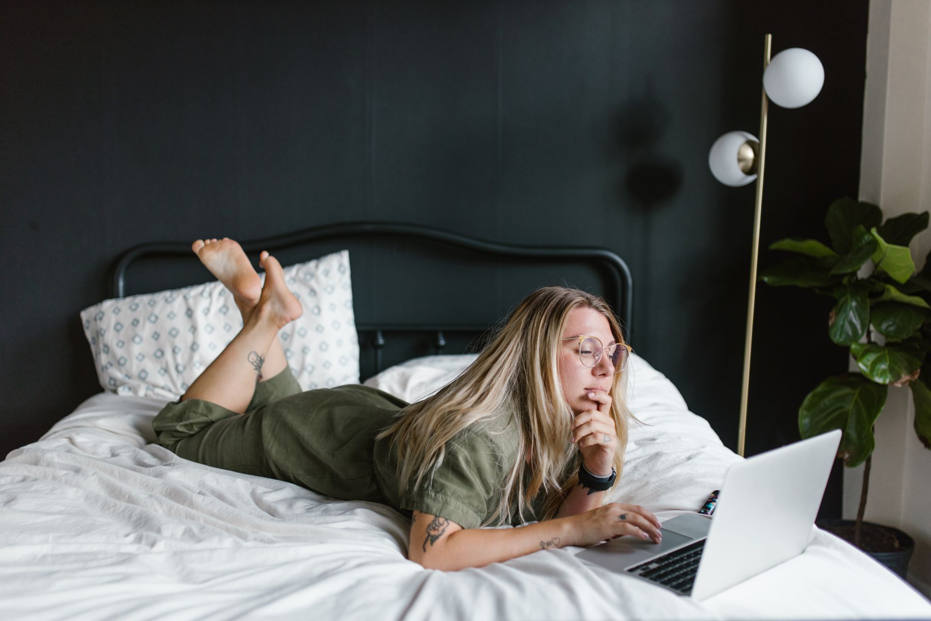 Woman in green dress using laptop on bed with floor lamp and plant.