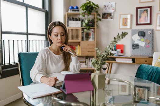 Asian woman working in a stylish home office, focusing on tasks at a glass table with tablet and papers.