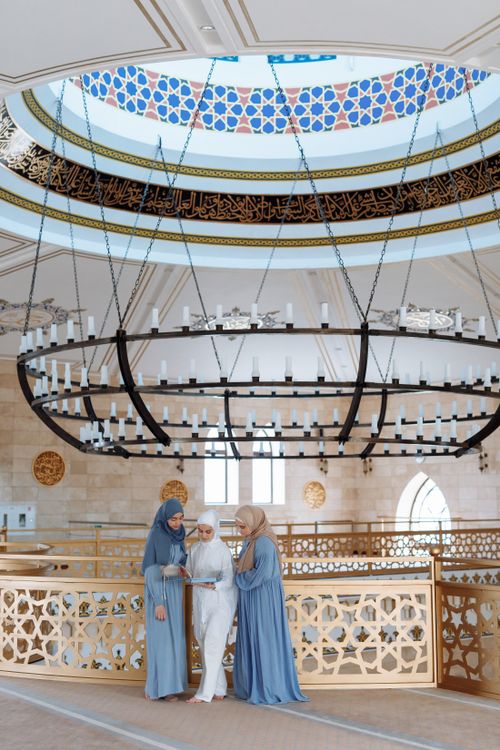 Free Three Muslim women in traditional attire read the Quran inside a grand mosque, emphasizing faith and togetherness. Stock Photo