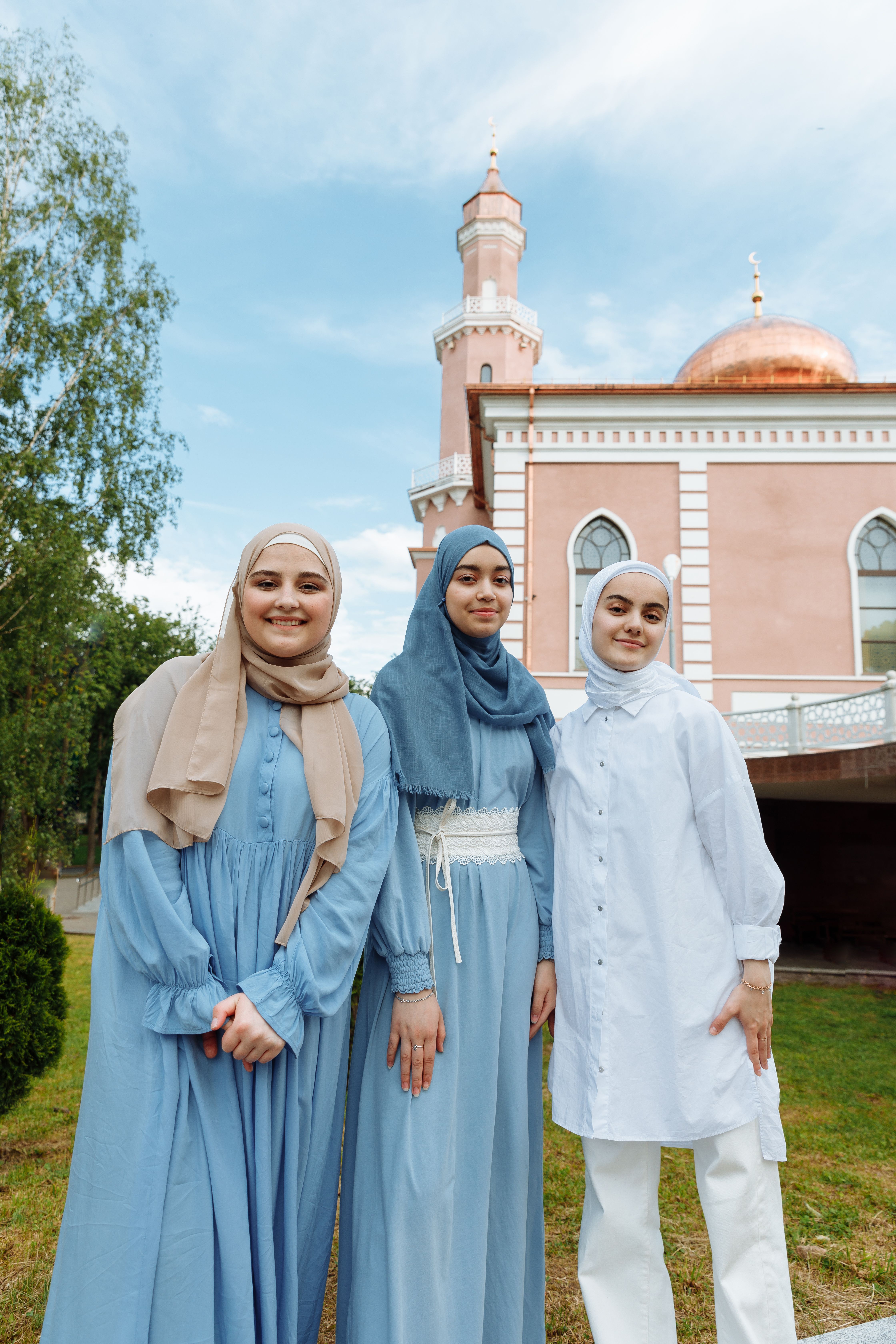 Three women wearing hijabs smiling in front of a mosque on a sunny day.