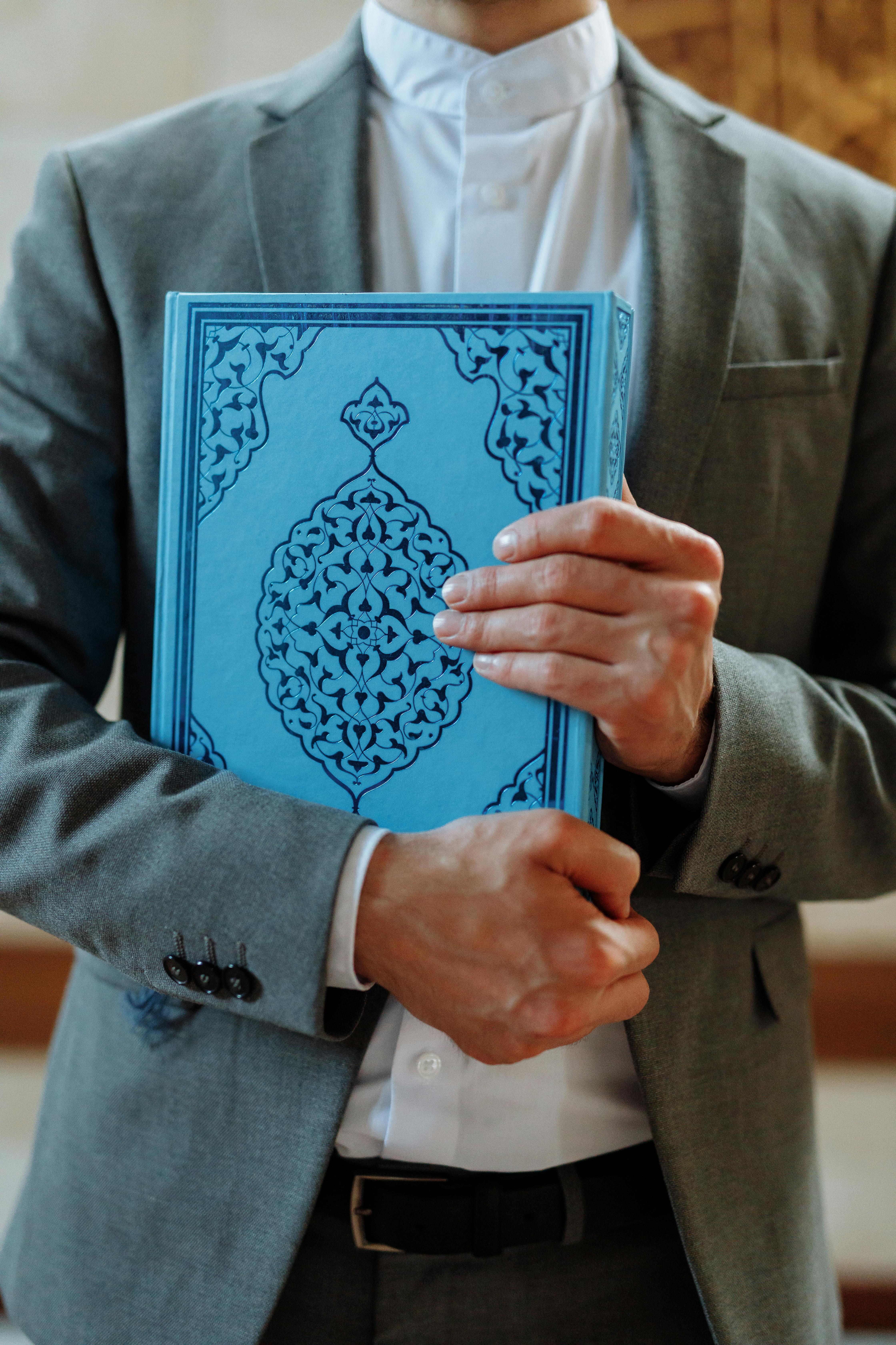 A person in a gray suit holds a blue Islamic book, symbolizing faith.