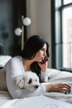 Woman multitasks on laptop and phone while relaxing with her dog in bed at home.