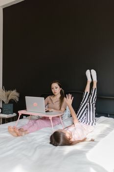 Mother working on laptop while daughter lies on bed in a cozy bedroom setting.