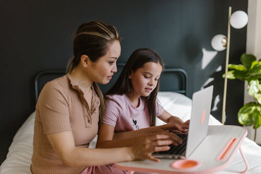 A mother and daughter sitting on a bed, using a laptop for learning together indoors.