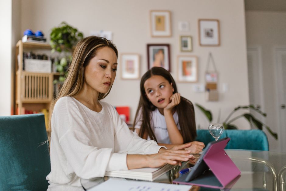 Una madre trabajando en su tableta en casa mientras su hija observa desde cerca, ilustrando los desafíos del teletrabajo.