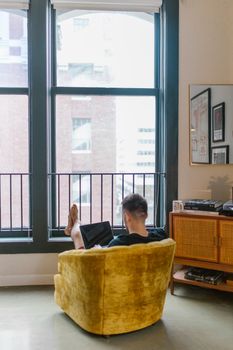 A man sits in a cozy home office looking at his laptop through large windows.
