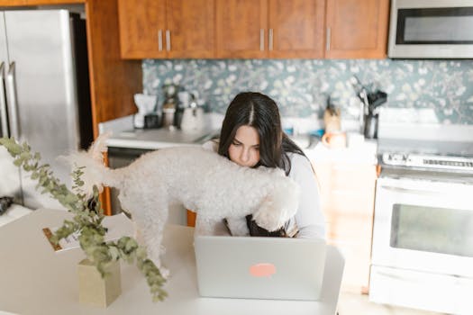 Woman working remotely on a laptop, accompanied by her pet dog in a modern kitchen setting.