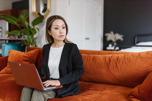 East Asian woman sitting on a sofa working from home with a laptop, wearing a stylish black blazer.