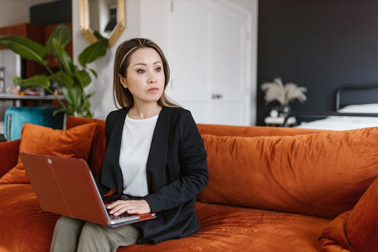 A Woman In Black Blazer Sitting On The Couch While Using Her Laptop