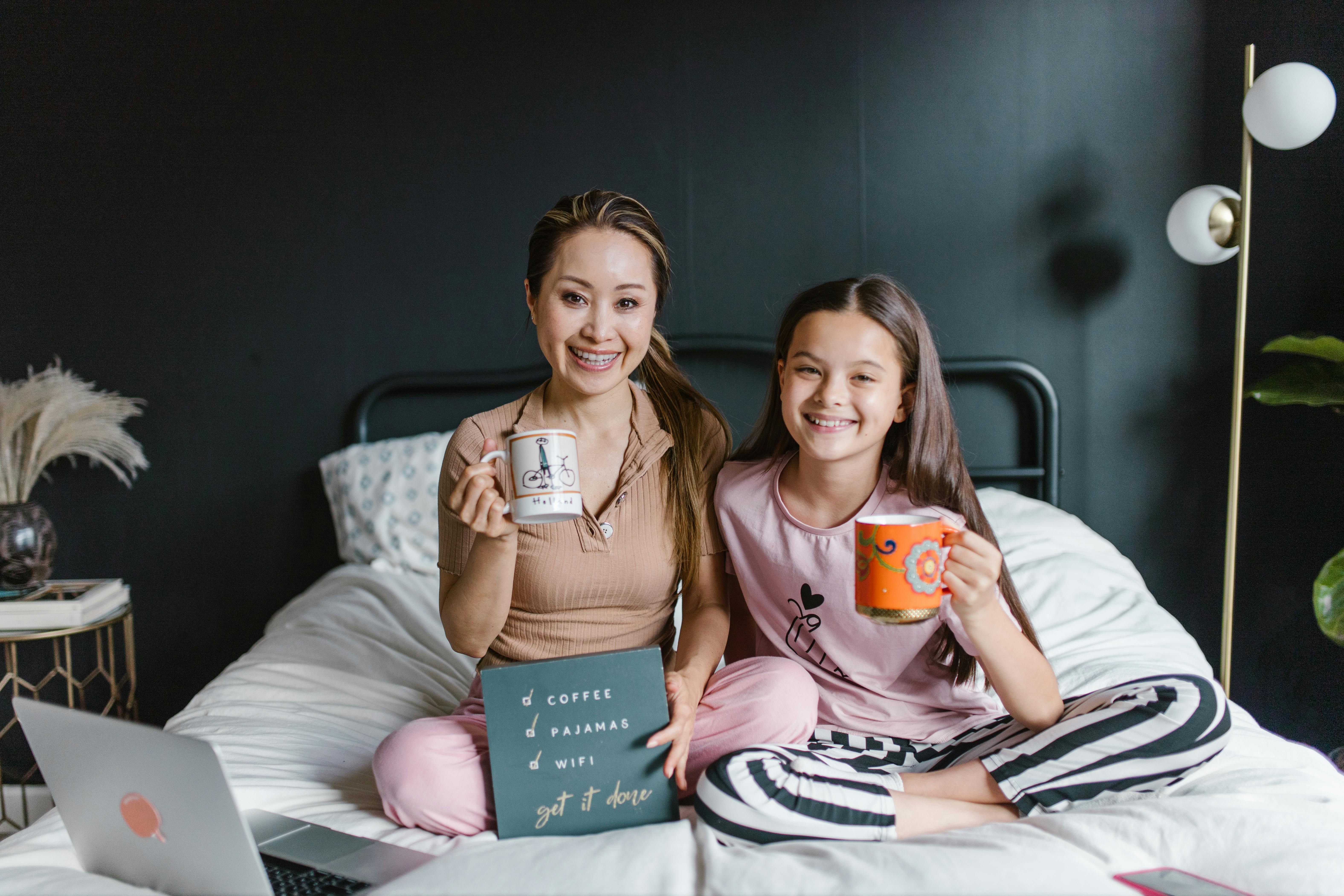 a mother and daughter sitting on the bed while holding a cup of coffee