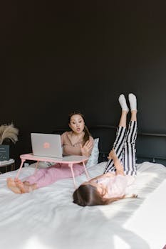 Woman using a laptop on bed while a child plays, embodying work-from-home balance.