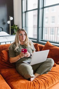 Woman with eyeglasses using smartphone and laptop on orange sofa in urban apartment.