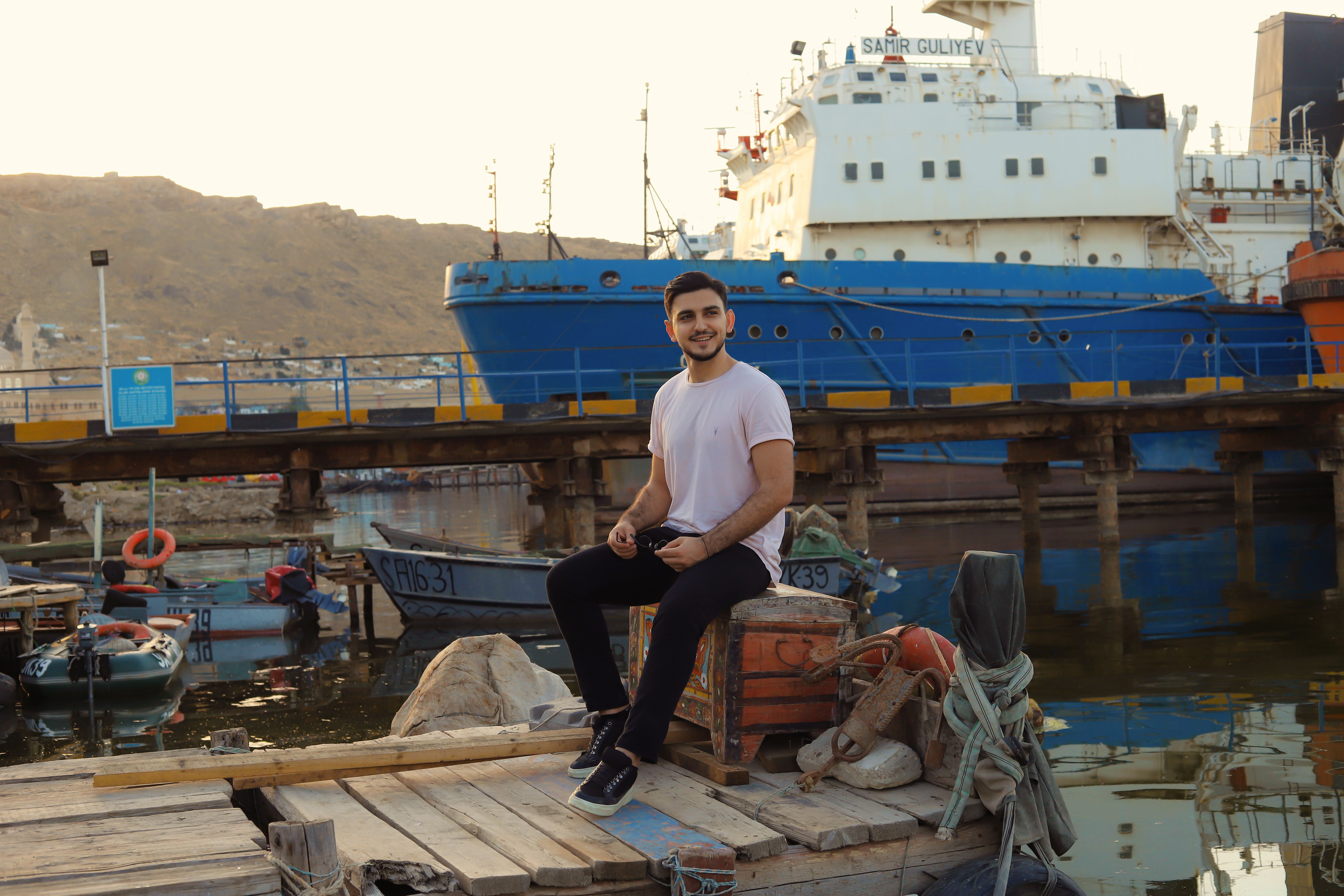 Young man sitting on a pier at a harbor with ships in the background under a clear sky.