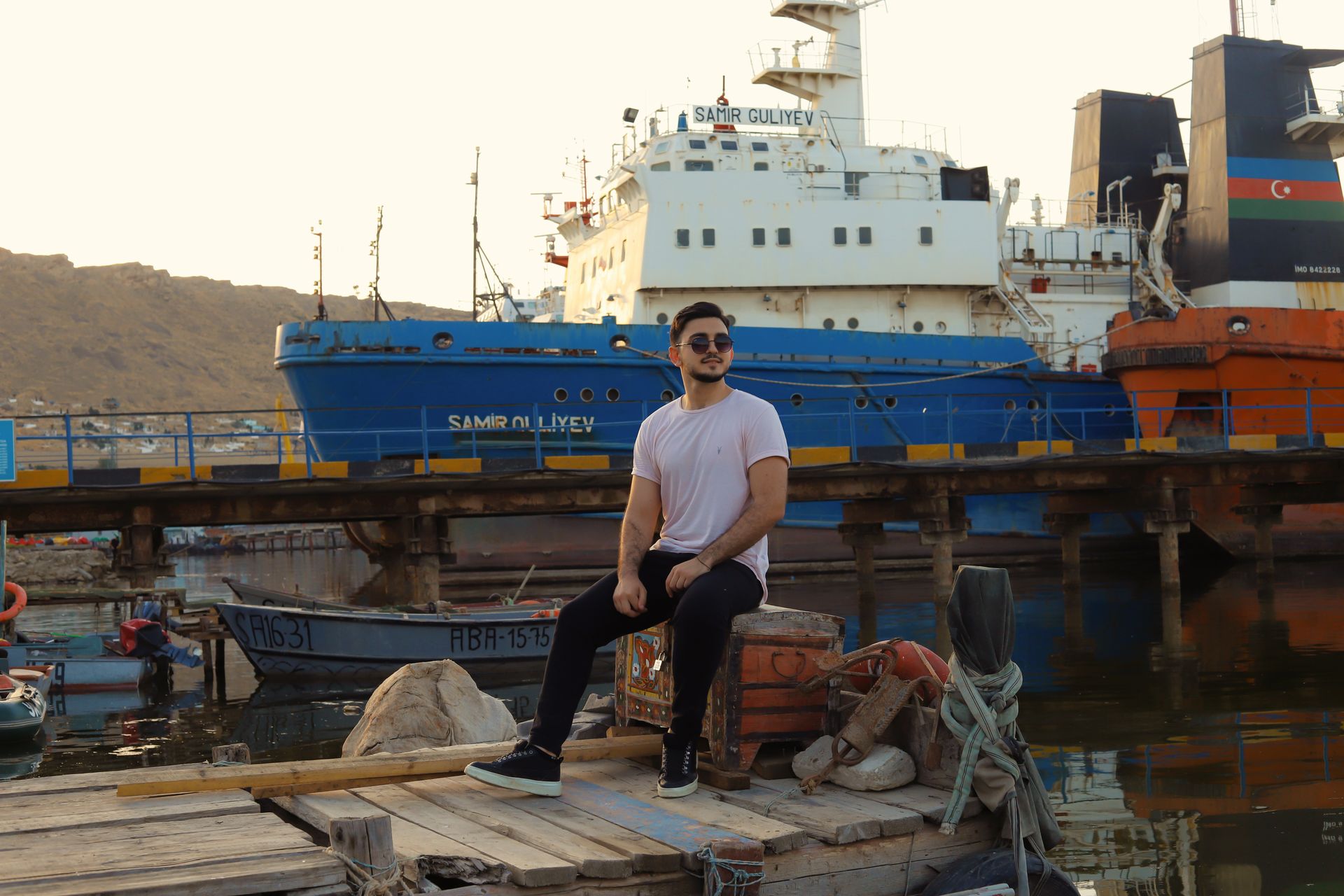 Portrait of a young man sitting on a dock with ships and containers at a bustling port.