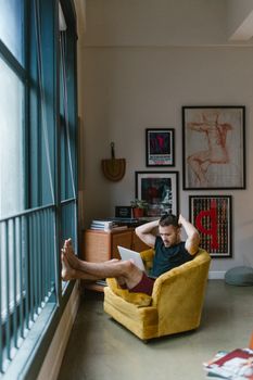 A man sitting comfortably in an armchair by the window, working on a laptop in a stylish home interior.