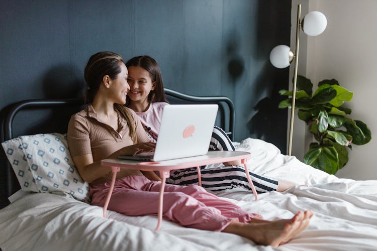 Young Women Smiling In Sleepwear Sitting On Bed Using A Computer Laptop