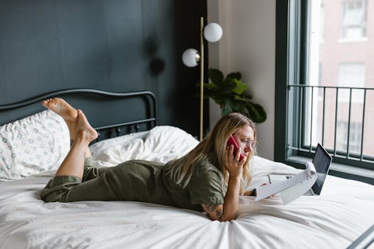 Woman on bed with laptop and phone, working remotely in cozy bedroom.