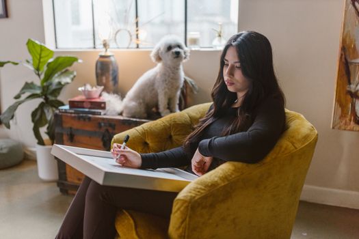 A woman artist sketching on a pad in a cozy room with a fluffy white dog by her side.