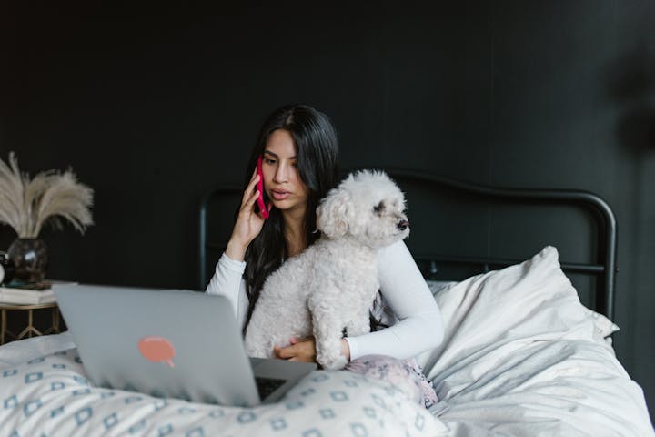 Woman talks on phone while working on laptop in bed with pet dog, cozy setting.