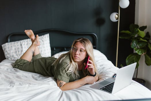 Woman using phone and laptop while working remotely from bed, surrounded by cozy decor.